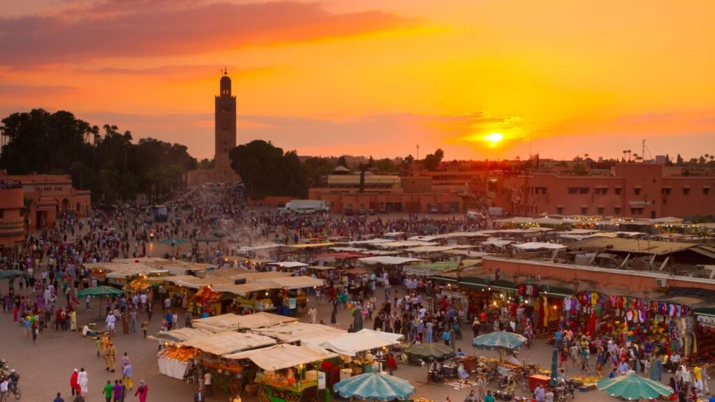 Atardecer en una plaza de Marrakech, el cielo está 100% naranja, hay tiendas, humo, gente caminando y comprando. A lo lejos se ve una torre, rodeada de árboles