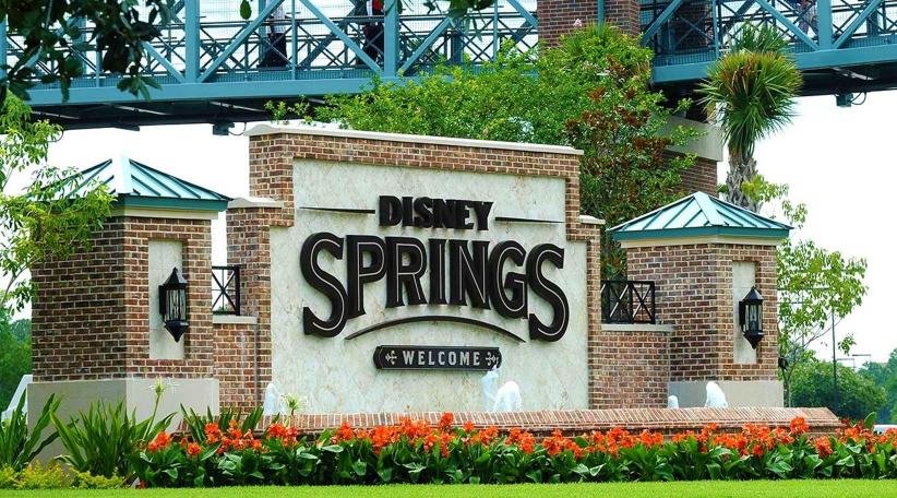 Cartel de Disney Springs, rodeado con un arco de ladrillos y flores rojas y pasto alrededor.