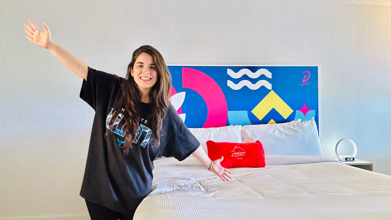 Juli sonriente posando en una habitación de hotel moderna y colorida, con una gran cabecera de cama decorada con formas geométricas y un cojín rojo que dice 'Developer Hotels'.
