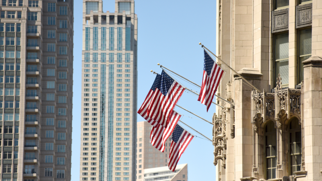 Edificios en NY, el cielo azul y dos banderas de EEUU asomando por el lado derecho