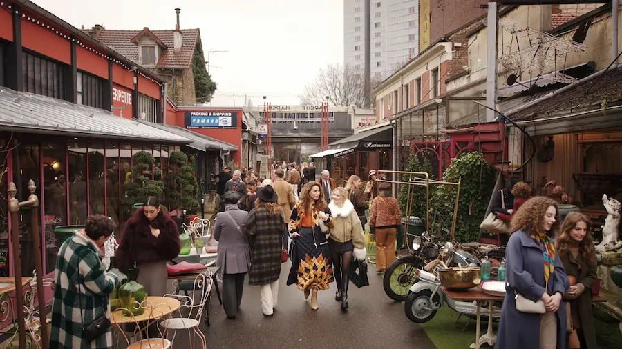 Mercado en una calle de Paris, el día está gris y dos personajes de la serie, Camille y su novia, caminan entre la gente.