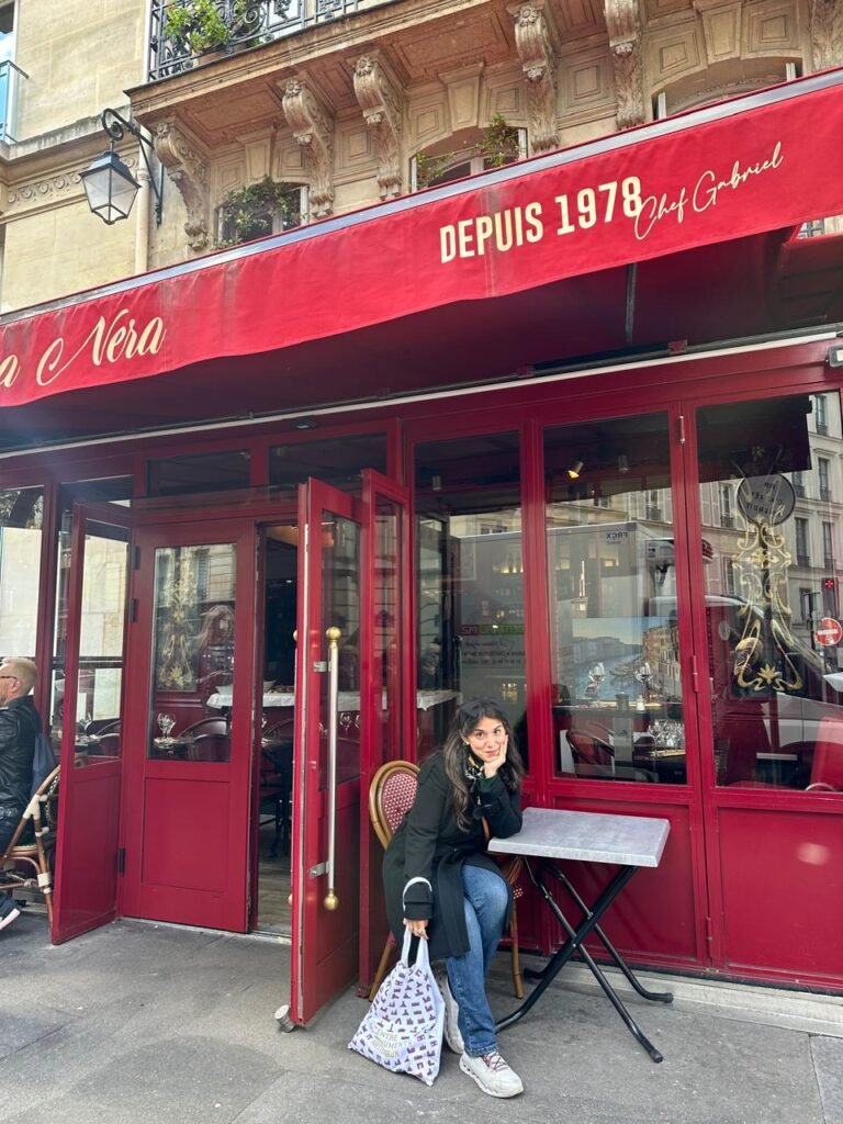 Juli sentada frente a la entrada del restaurante Casa Nera en París, con un toldo rojo que muestra ‘Desde 1978 Chef Gabriel’ y el ambiente acogedor de una típica cafetería parisina.