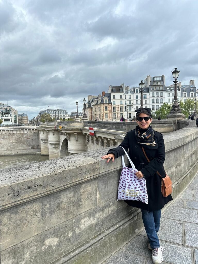 Juli con abrigo negro y bufanda posando junto al Puente Neuf en París, con el río Sena y edificios históricos bajo un cielo nublado de fondo.