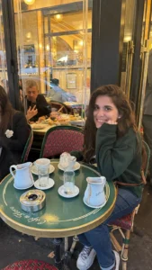 Juli sonriendo a cámara sentada en una de las mesas verdes del Cafe di flore, con dos chocolates caliente en loza blancas.