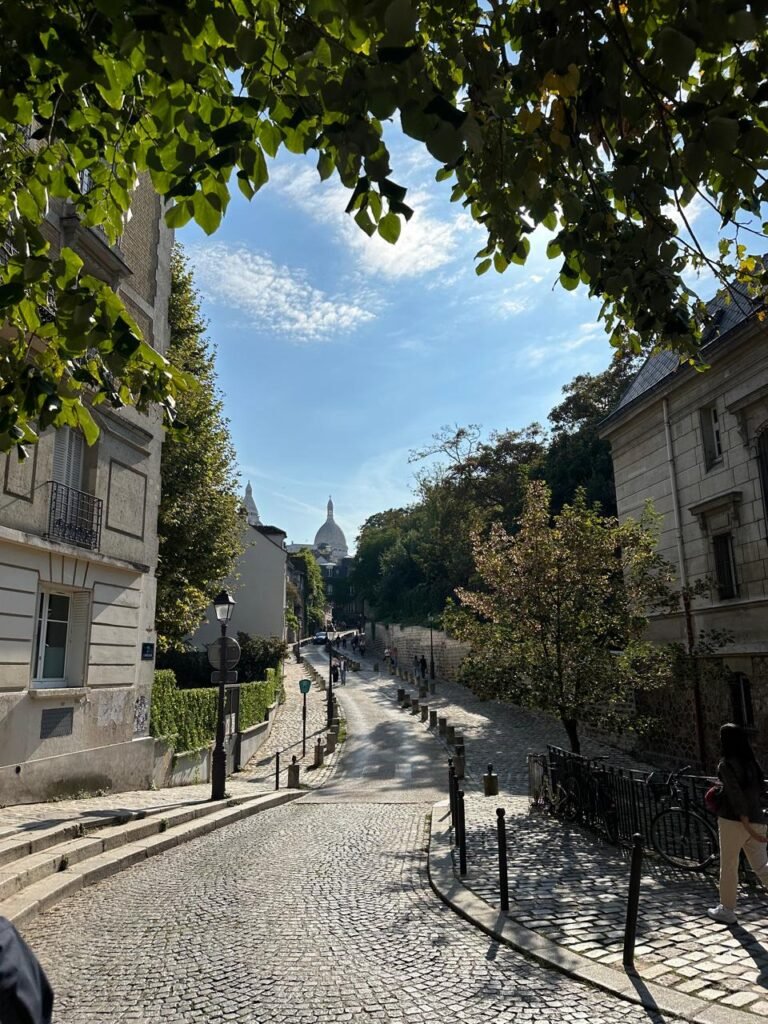Vista de una calle empedrada en Montmartre, que lleva hacia la icónica Basílica del Sacré-Cœur, enmarcada por árboles y edificios históricos. La luz del sol atravesando las hojas añade un toque mágico a esta clásica escena parisina.