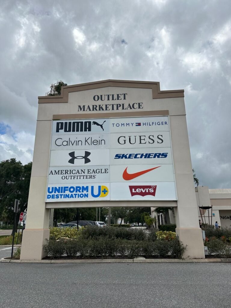 Cartel del Outlet Marketplace en Orlando, Florida, mostrando las principales marcas disponibles en este centro comercial, como Puma, Calvin Klein, Tommy Hilfiger, Guess, American Eagle Outfitters, Skechers, Under Armour, Nike, Levi's y Uniform Destination. El cielo parcialmente nublado crea un ambiente típico de la región.