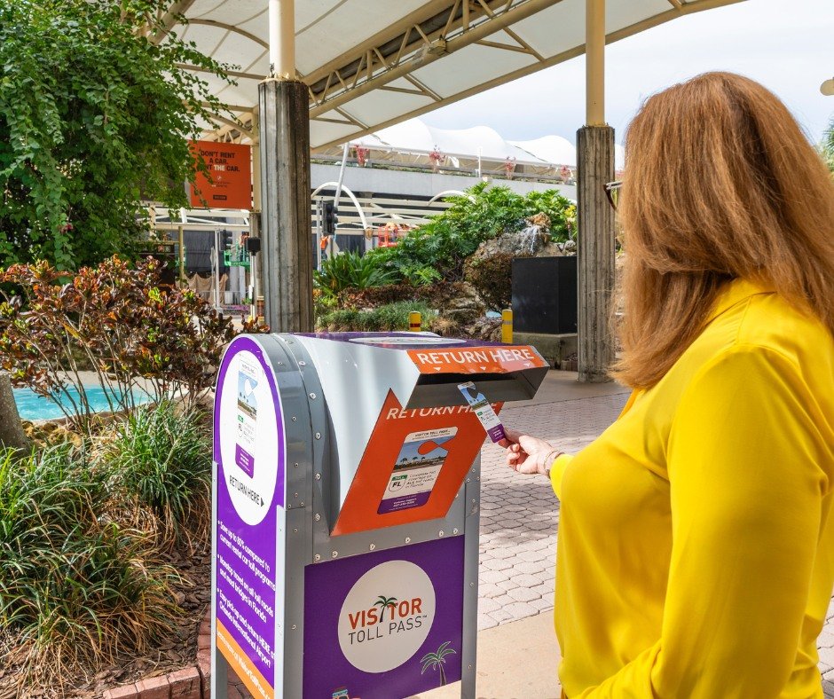 Mujer de cabello castaño claro y blusa amarilla devolviendo un Visitor Toll Pass en un buzón de devolución ubicado en un área exterior cubierta, rodeada de plantas y señalizaciones.