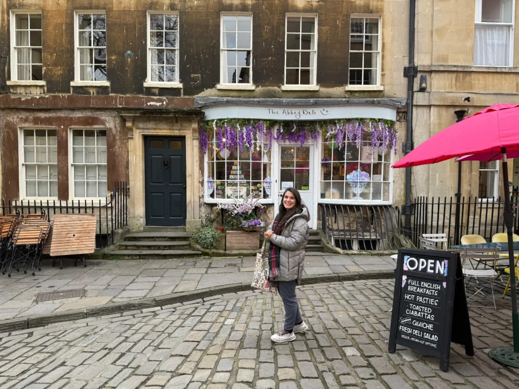 Juli sonriente frente a The Abbey Deli, una pintoresca cafetería con flores colgantes en la fachada, ubicada en una calle empedrada de Bath, Inglaterra. El cartel indica que el lugar ofrece desayunos ingleses, pasteles calientes, ciabattas, quiches y ensaladas frescas, con opciones veganas.