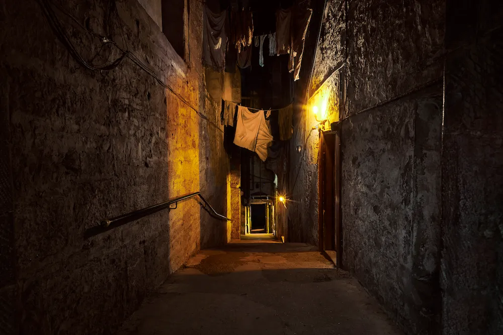 Callejón angosto de noche con paredes de piedra y luz cálida de farol, ropa colgando en lo alto entre los edificios.
