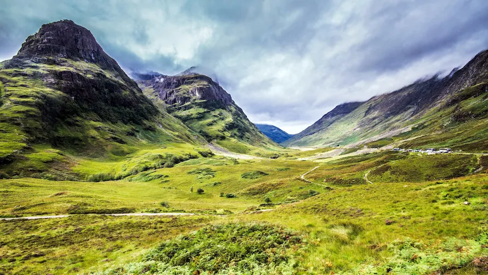 Valle verde rodeado de montañas escarpadas en Glencoe, Escocia. El cielo está parcialmente cubierto de nubes grises, mientras senderos serpentean entre la vegetación, evocando un paisaje dramático y natural.