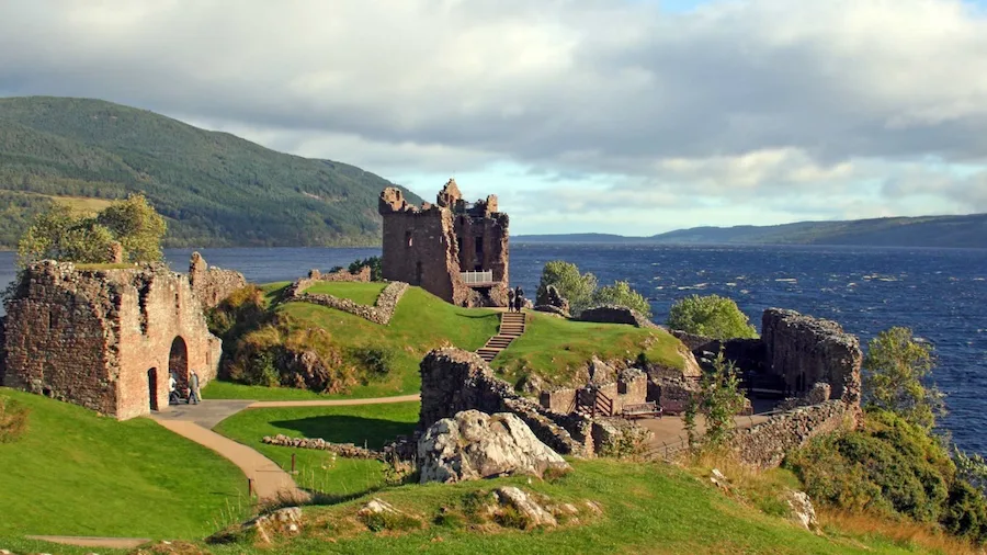 Ruinas del castillo de Urquhart sobre una colina verde, con vista al lago Ness en Escocia. El castillo de piedra parcialmente derruido se encuentra junto al agua, rodeado de árboles y caminos, bajo un cielo parcialmente nublado.