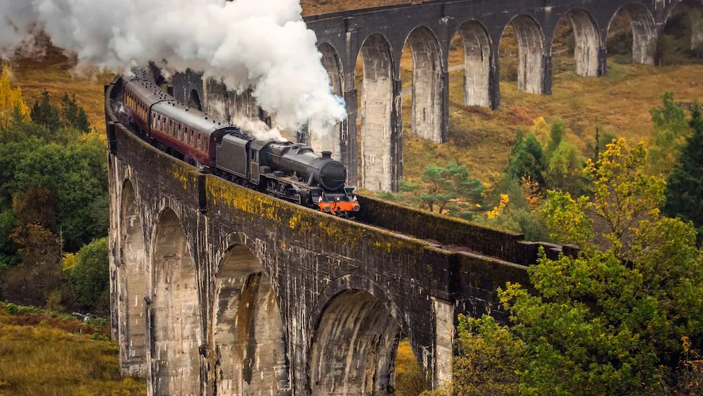 Tren a vapor cruzando el viaducto de Glenfinnan en Escocia, con columnas de piedra y paisaje otoñal de fondo. El tren emite una gran nube de vapor blanco mientras atraviesa el puente curvo sobre un valle rodeado de árboles.