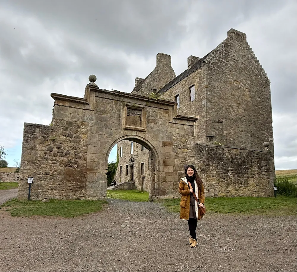 Juli frente a la entrada arqueada de un castillo antiguo de piedra, en un día nublado. El edificio tiene muros altos, ventanas pequeñas y un estilo medieval. Ella viste abrigo marrón claro y sonríe hacia la cámara.