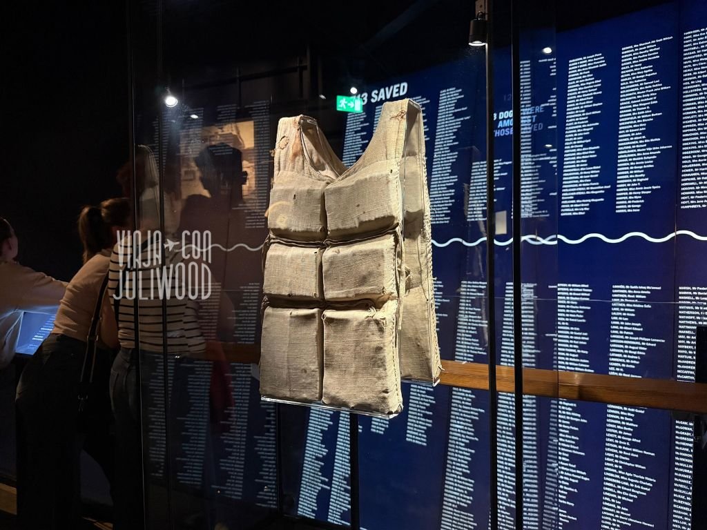 Chaleco salvavidas original del Titanic exhibido en vitrina de cristal frente a una pared con los nombres de los pasajeros.