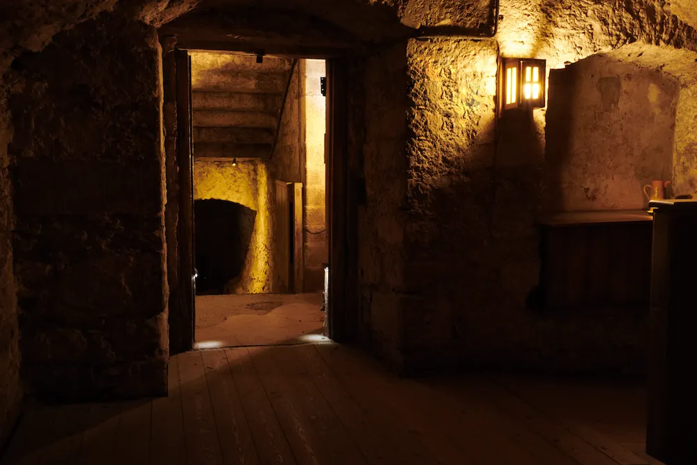 Interior de piedra iluminado tenuemente con luz cálida, mostrando una puerta abierta hacia una escalera de piedra antigua y una pared curva en penumbra.