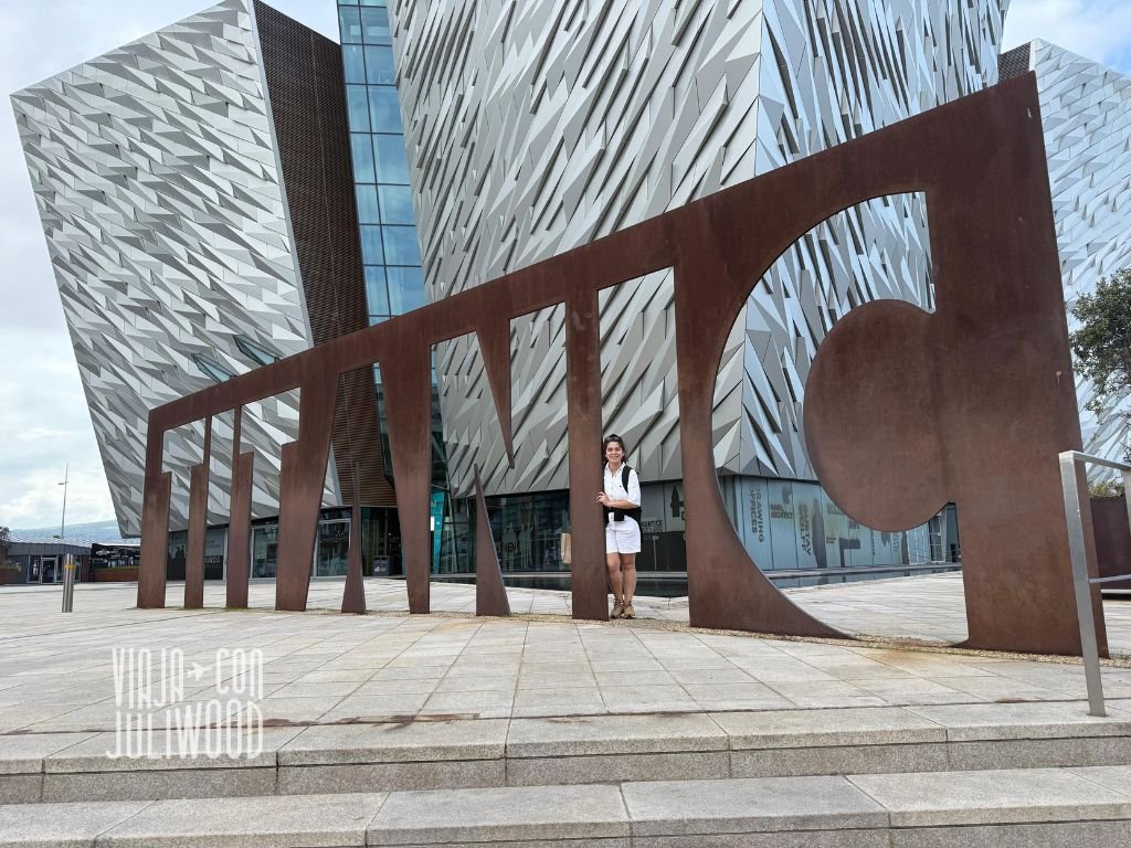 Mujer posando junto a la escultura de metal con la palabra “TITANIC” frente al icónico edificio del museo Titanic Belfast.