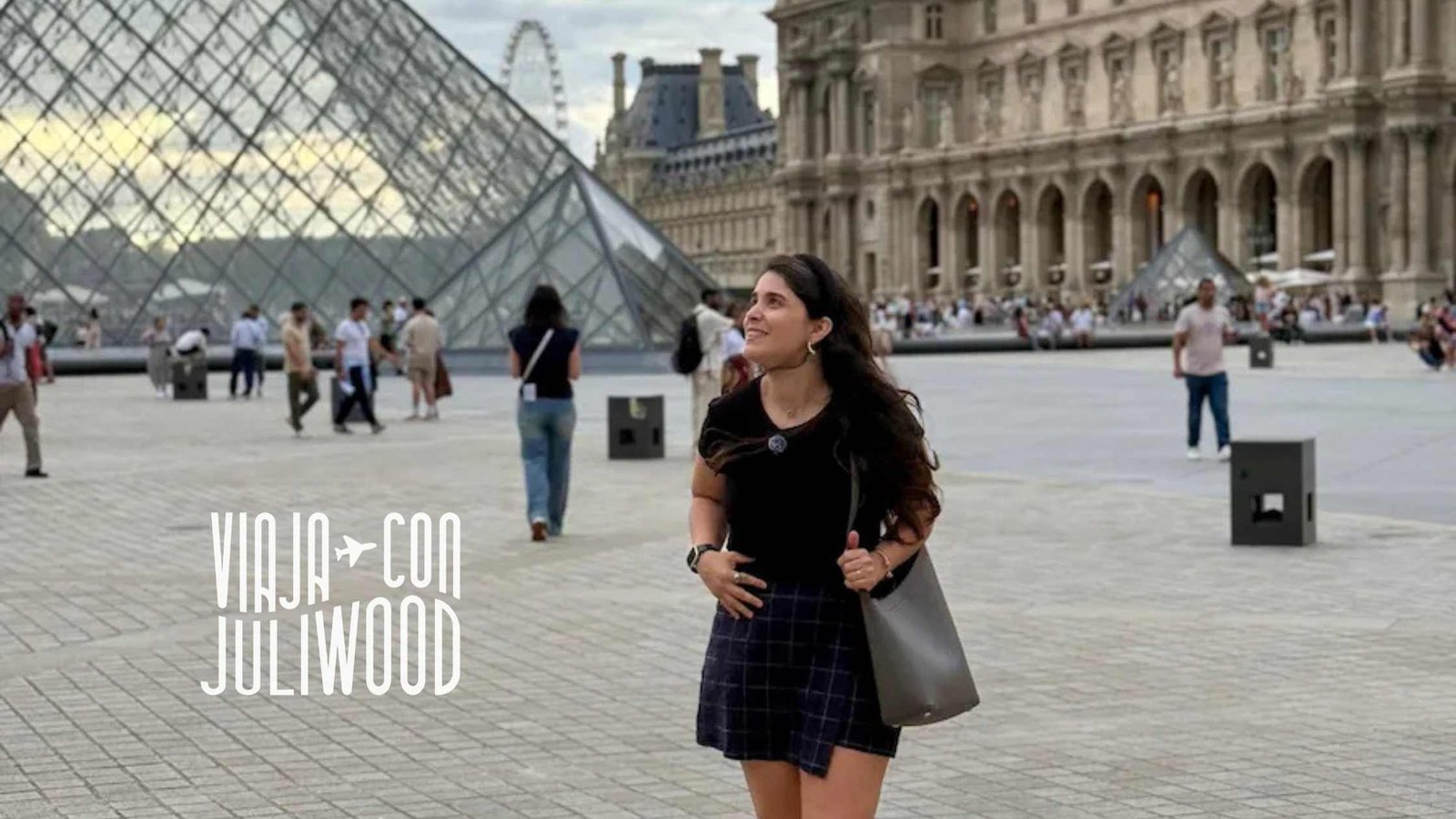 sonriendo mientras camina frente a la Pirámide de Cristal del Museo del Louvre en París, con arquitectura clásica de fondo y turistas alrededor.