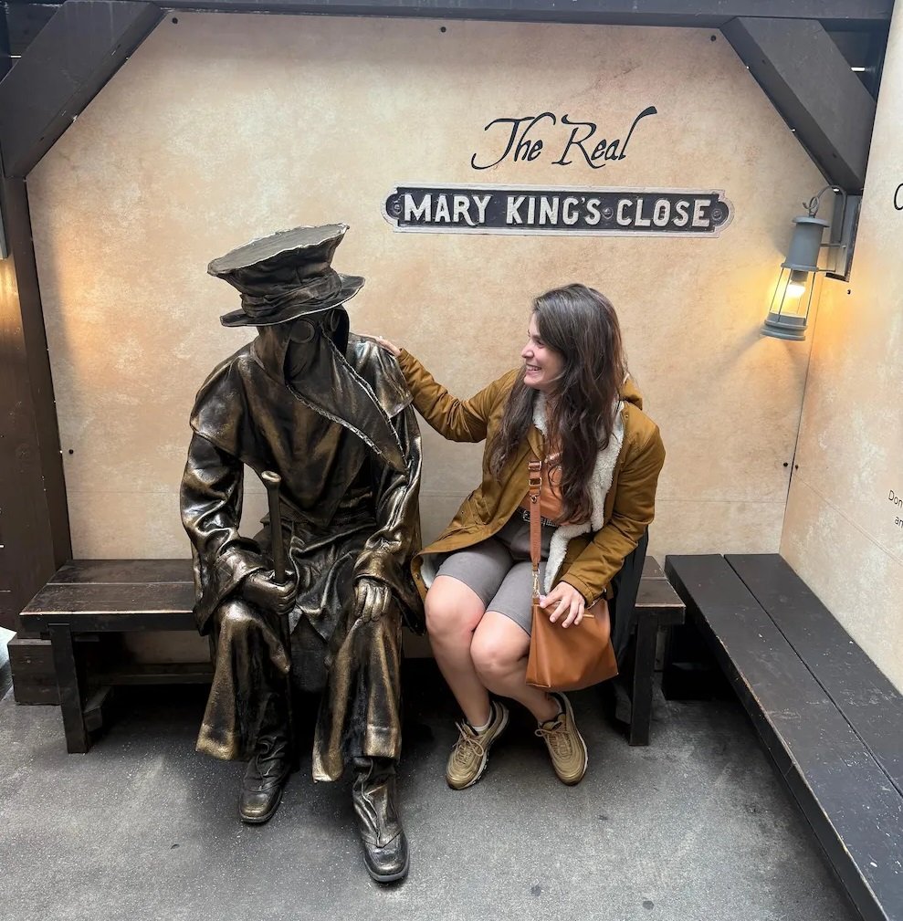 Juli con una estatua de la figura de médico de la peste con máscara de pico, vestido con túnica marrón oscura y bastón.
