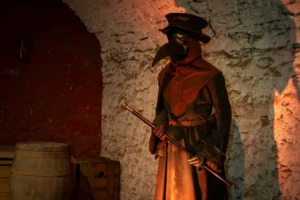 Figura de médico de la peste con máscara de pico, vestido con túnica marrón oscura y bastón, en una habitación de piedra con iluminación tenue.