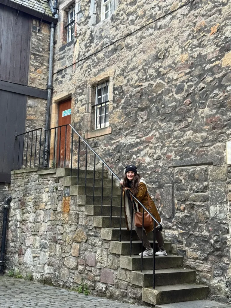 Juli sonriendo sobre una escalera de piedra junto a una antigua pared de ladrillos en un callejón empedrado de Edimburgo, con abrigo y gorro de invierno.