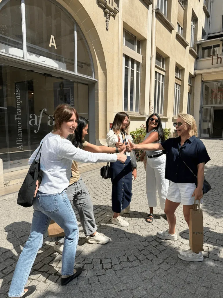 Grupo de amigas riendo y jugando a piedra, papel o tijera frente a la entrada principal de la Alliance Française de París. El edificio de estilo clásico se ve al fondo, con puertas de vidrio y el logo "af" visible. El clima es soleado y relajado, con un ambiente de despedida o cierre de jornada. Una de ellas sostiene una bolsa de papel en la mano, otra lleva gafas de sol, y todas comparten un momento de diversión espontánea.