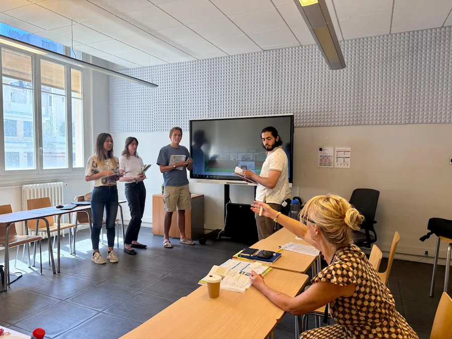 Estudiantes realizando una actividad oral frente a la clase con libros en mano, mientras la docente, sentada en el escritorio, les indica algo con el brazo extendido. Aula luminosa y moderna.