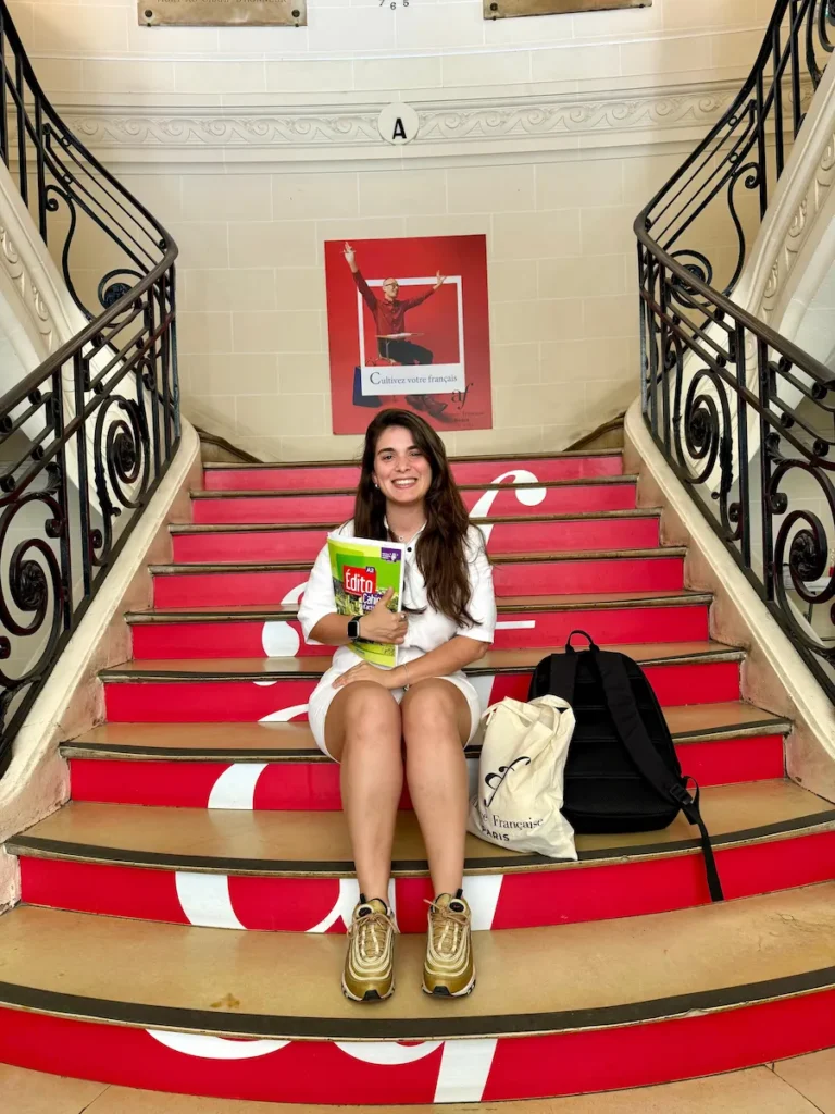 Juli sentada en una escalera decorada con letras rojas en el interior de la Alliance Française de París. Sostiene libros de estudio “Édito A2” y una bolsa con el logo de la institución, sonriendo a cámara.