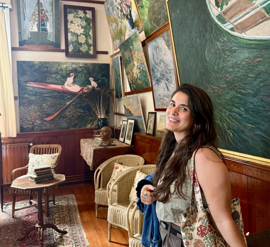 Juli sonriendo dentro de una sala decorada con cuadros inspirados en el impresionismo francés, posiblemente de Monet. El ambiente es cálido, con sillones de mimbre y libros antiguos.