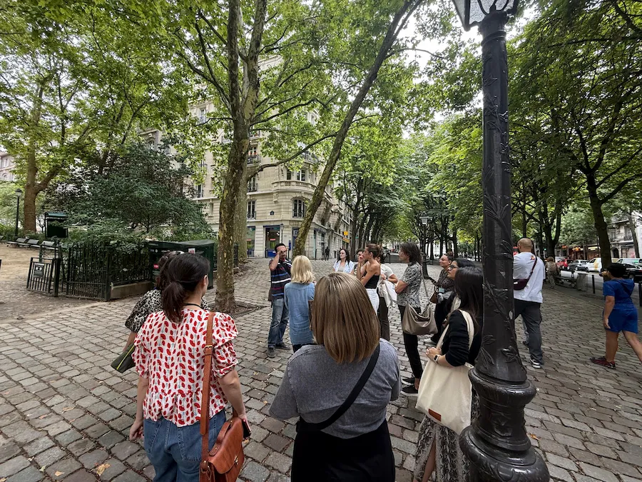 Grupo de personas participando en una visita guiada por una calle empedrada y arbolada de París. Un guía habla mientras los participantes observan atentos los alrededores.