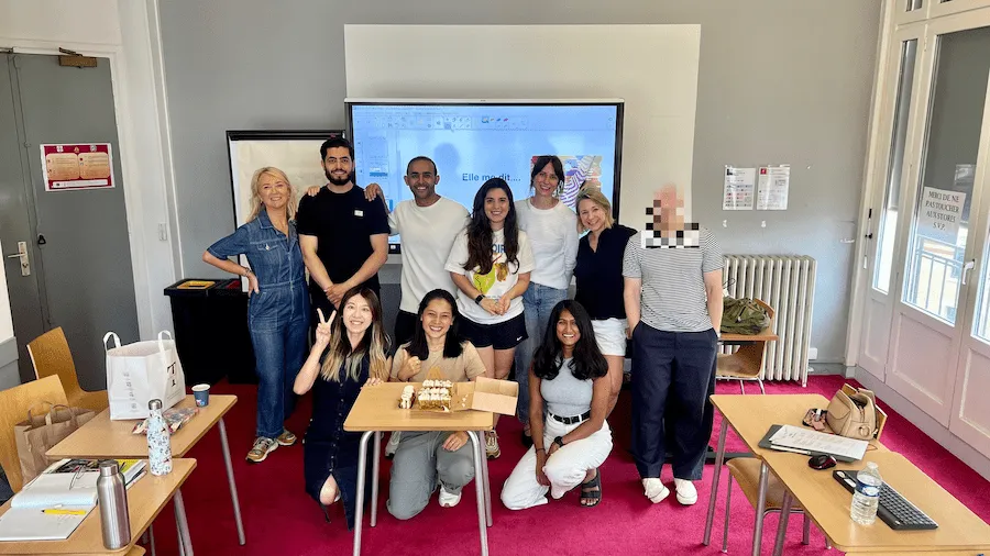 Grupo de estudiantes posando dentro de un aula con una profesora, frente a una pantalla que muestra la frase “Elle m’a dit…”. Hay escritorios con libros, agua y torta. Todos sonríen, celebrando posiblemente el cierre de curso.