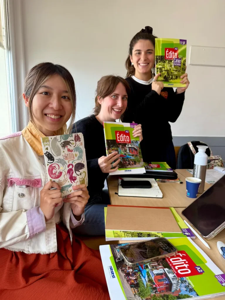 Tres estudiantes sentadas en clase mostrando sus cuadernos y libros del curso de francés “Édito A2”. Se ven sonrisas, materiales de estudio y un ambiente cálido y colaborativo.