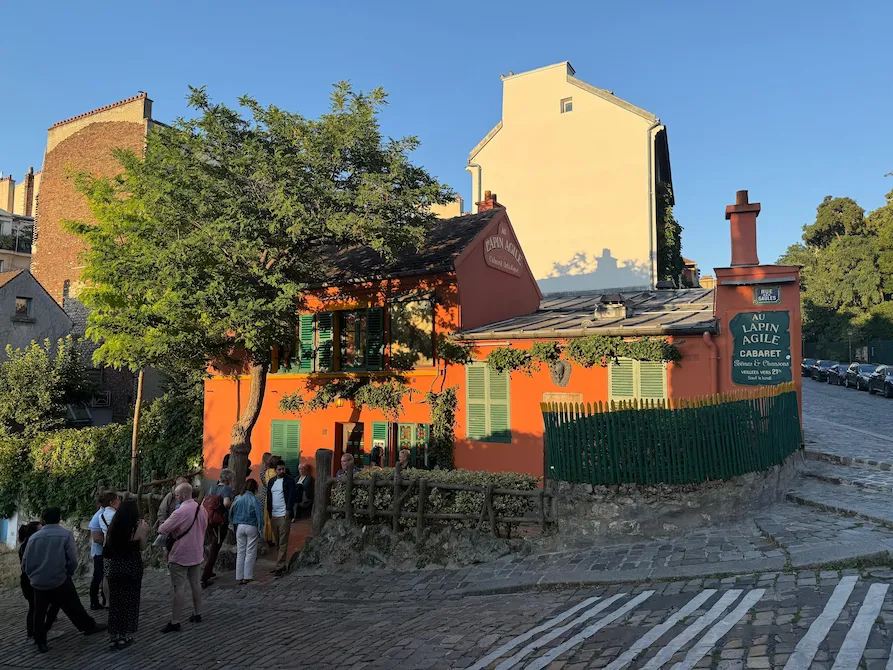 Fachada del cabaret Le Lapin Agile en Montmartre, París, al atardecer, con visitantes reunidos frente a la casa color terracota.