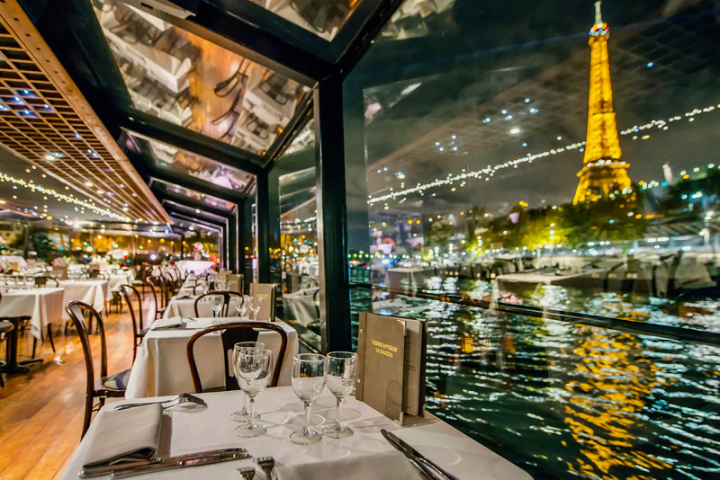 Cena a bordo de un crucero por el Sena en París, con mesas elegantes y vista nocturna de la Torre Eiffel iluminada reflejándose en el río.