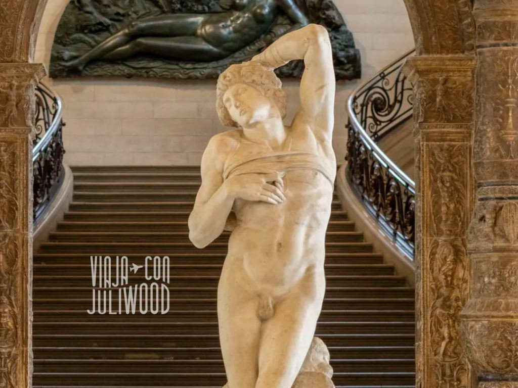 Escultura del Esclavo Rebelde de Miguel Ángel en el Louvre, figura masculina desnuda en tensión, ubicada frente a una escalera monumental.