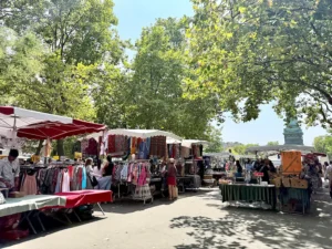 Puestos del mercado al aire libre en París con ropa y accesorios, rodeados de árboles y visitantes recorriendo las paradas en un día soleado.