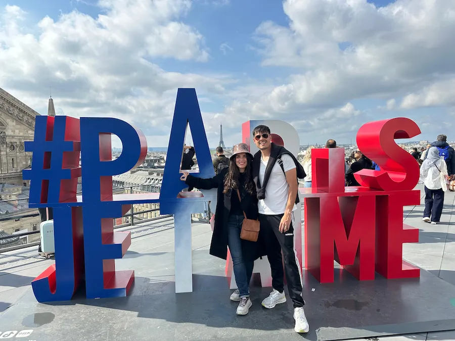 Pareja posando frente a las letras gigantes “PARIS JE T’AIME” en la terraza de Galeries Lafayette, con la Torre Eiffel al fondo.