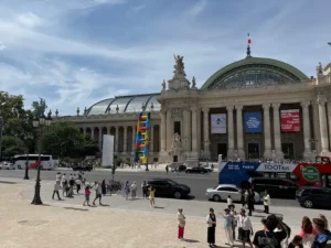 Fachada del Grand Palais en París en un día soleado, con su cúpula de vidrio, esculturas clásicas y una colorida instalación vertical frente al edificio.