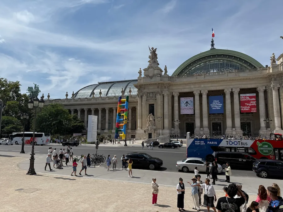 Fachada del Grand Palais en París en un día soleado, con su cúpula de vidrio, esculturas clásicas y una colorida instalación vertical frente al edificio.