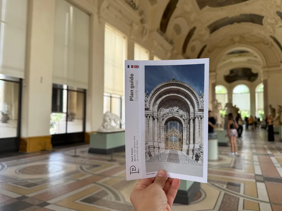 Mano sosteniendo el plano del Petit Palais dentro de una sala amplia con esculturas, ventanales altos y suelos decorados.