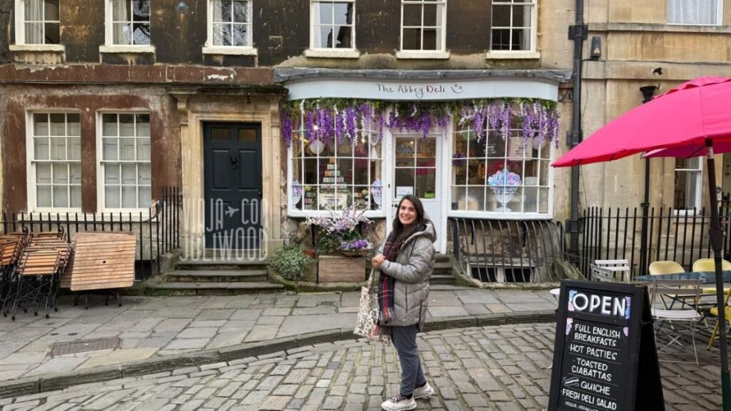 Plazoleta Abbey Green en Bath, con suelo empedrado, fachadas históricas y la tienda The Abbey Deli decorada con flores violetas; Juli está de pie sonriendo frente al local.