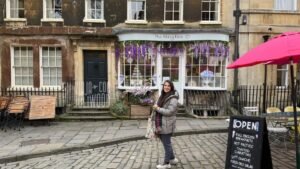 Plazoleta Abbey Green en Bath, con suelo empedrado, fachadas históricas y la tienda The Abbey Deli decorada con flores violetas; Juli está de pie sonriendo frente al local.