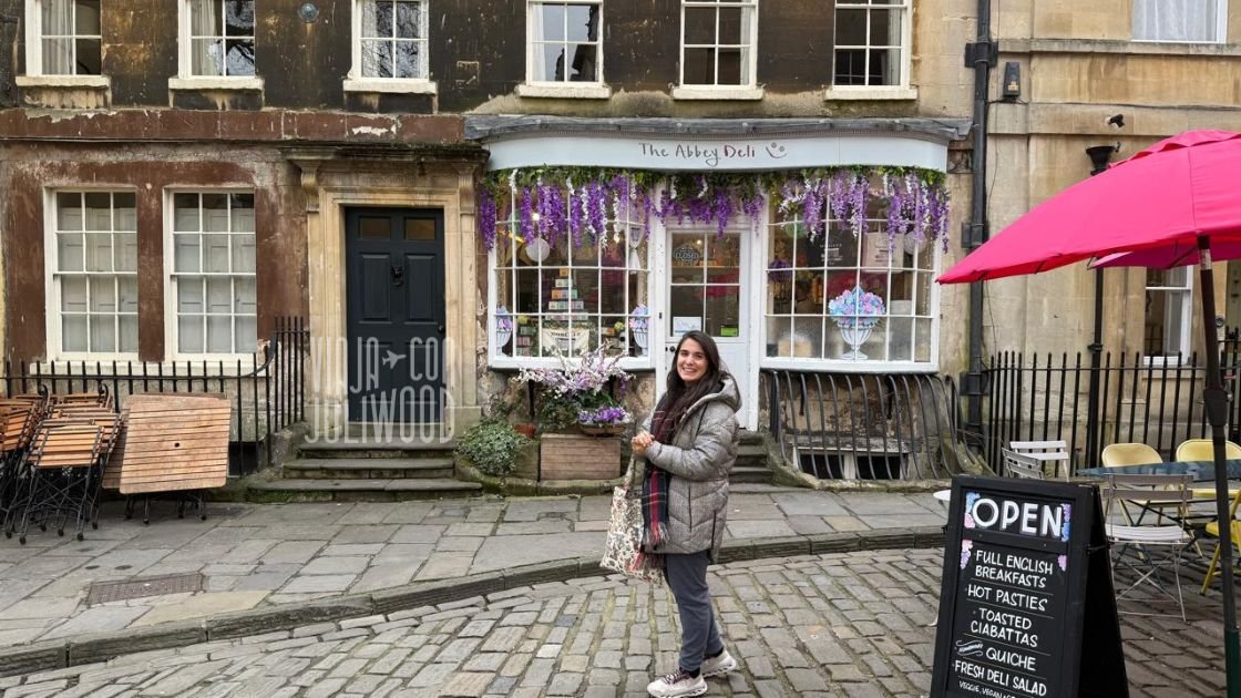 Plazoleta Abbey Green en Bath, con suelo empedrado, fachadas históricas y la tienda The Abbey Deli decorada con flores violetas; Juli está de pie sonriendo frente al local.