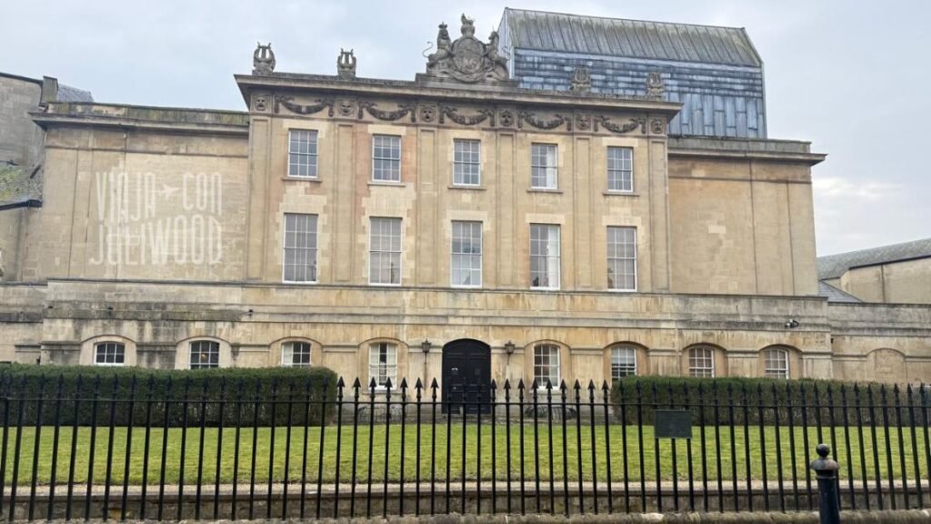 Edificio histórico de piedra color beige en Bath, con fachada simétrica de varios pisos, ventanas rectangulares alineadas y un pequeño jardín delantero cercado con rejas negras.
