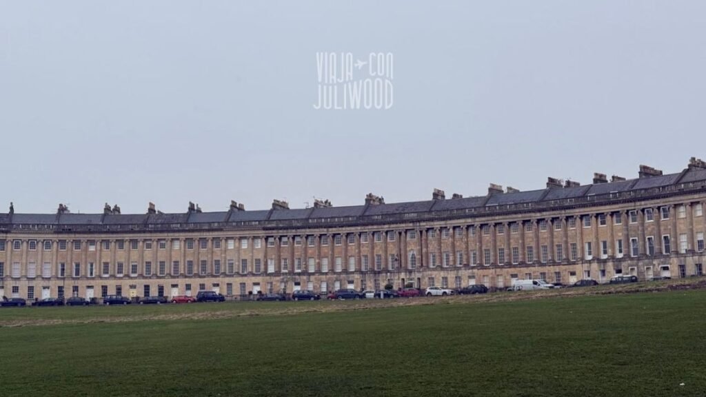 Vista amplia del Royal Crescent en Bath, una larga fila curva de casas georgianas de piedra clara, frente a un gran parque verde con césped.
