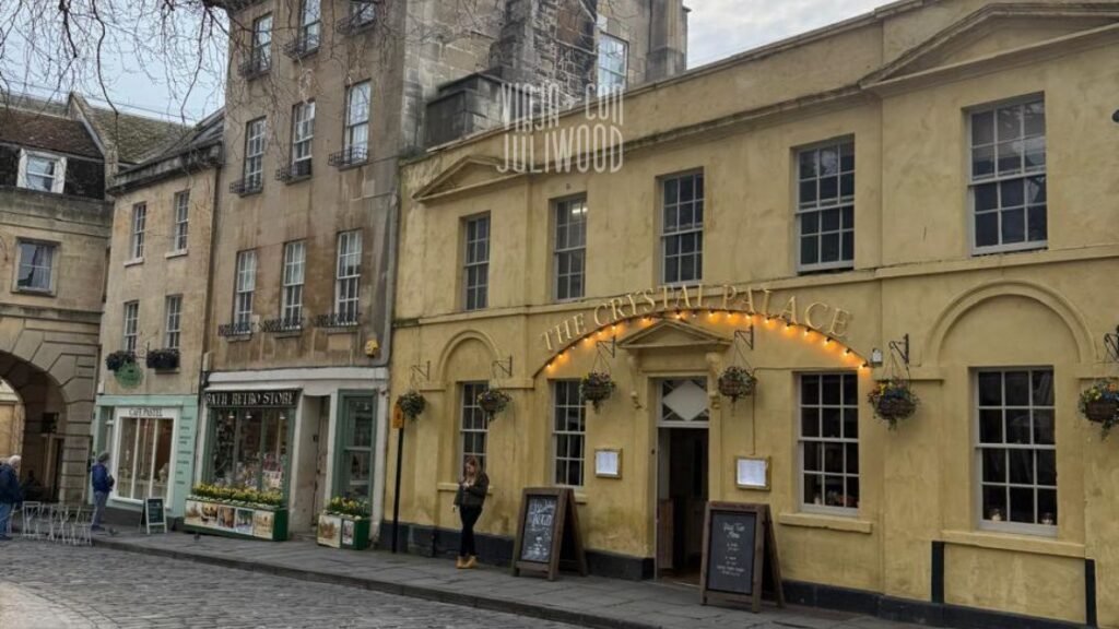 Calle empedrada en el centro de Bath con edificios antiguos de piedra; a la derecha se ve un pub de fachada amarilla llamado The Crystal Palace, con luces cálidas y mesas en la vereda.