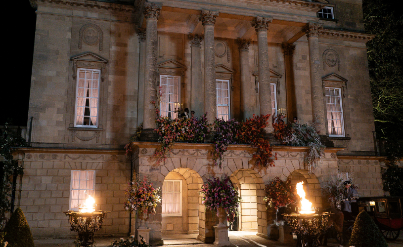 Edificio histórico iluminado de noche, con columnas y balcones decorados con flores, antorchas encendidas a ambos lados de la entrada y personas vestidas de época, escena de una filmación.