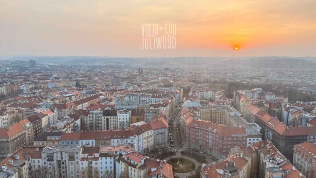 Vista panorámica de Praga al atardecer desde lo alto, con techos rojos y el sol sobre la ciudad.